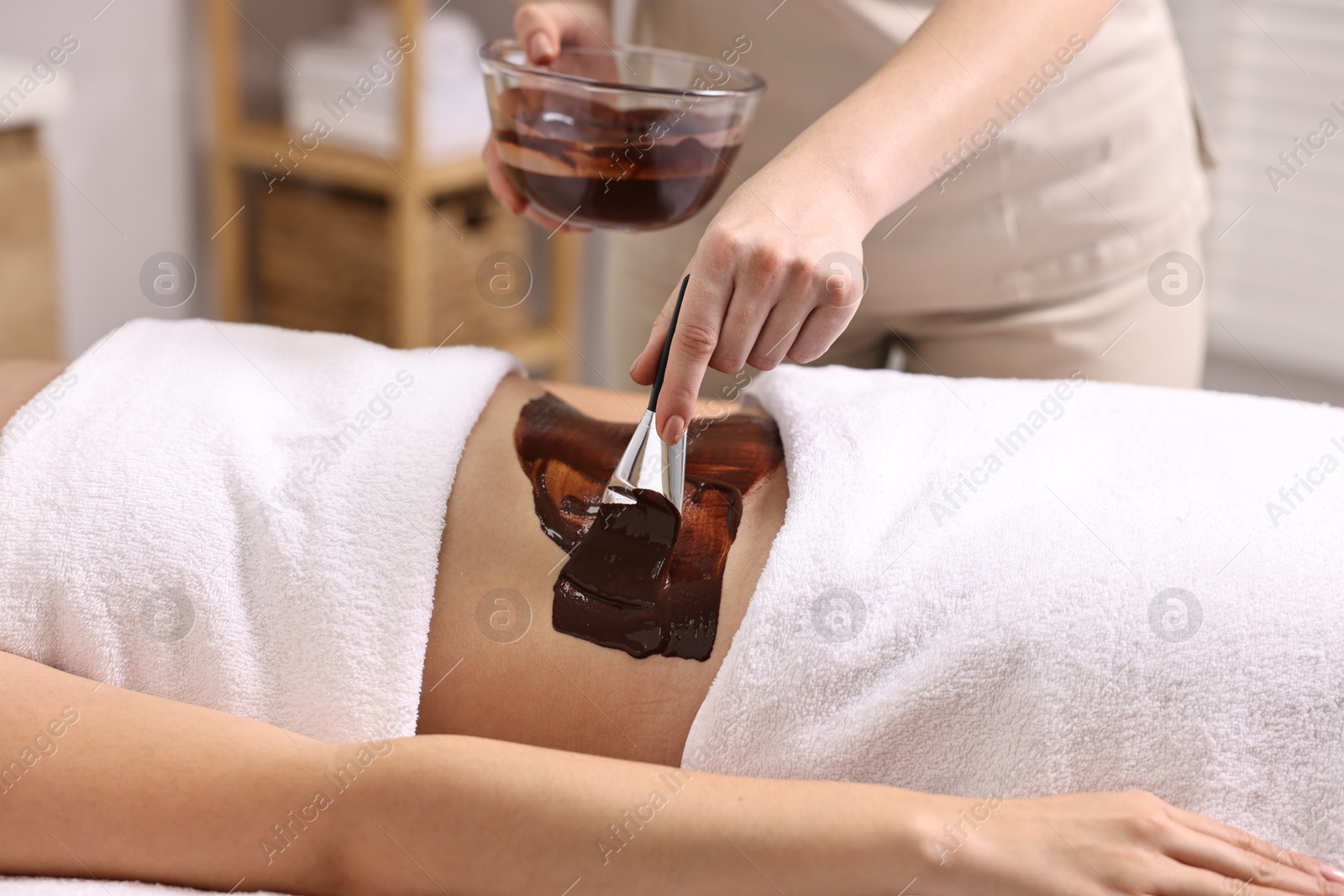 Photo of Chocolate body wrap. Spa worker applying mask onto woman's belly in salon, closeup