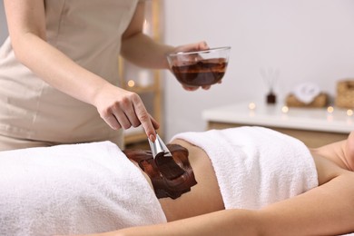 Photo of Chocolate body wrap. Spa worker applying mask onto woman's belly in salon, closeup