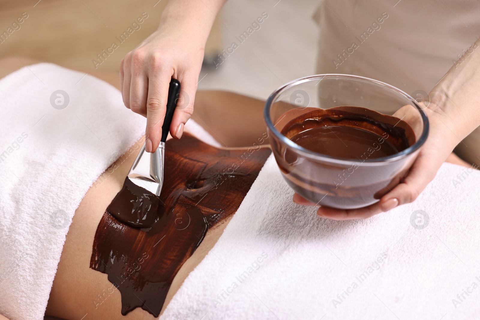 Photo of Chocolate body wrap. Spa worker applying mask onto woman's belly in salon, closeup