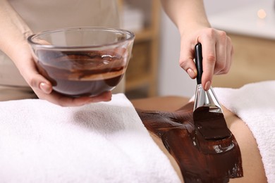 Photo of Chocolate body wrap. Spa worker applying mask onto woman's belly in salon, closeup