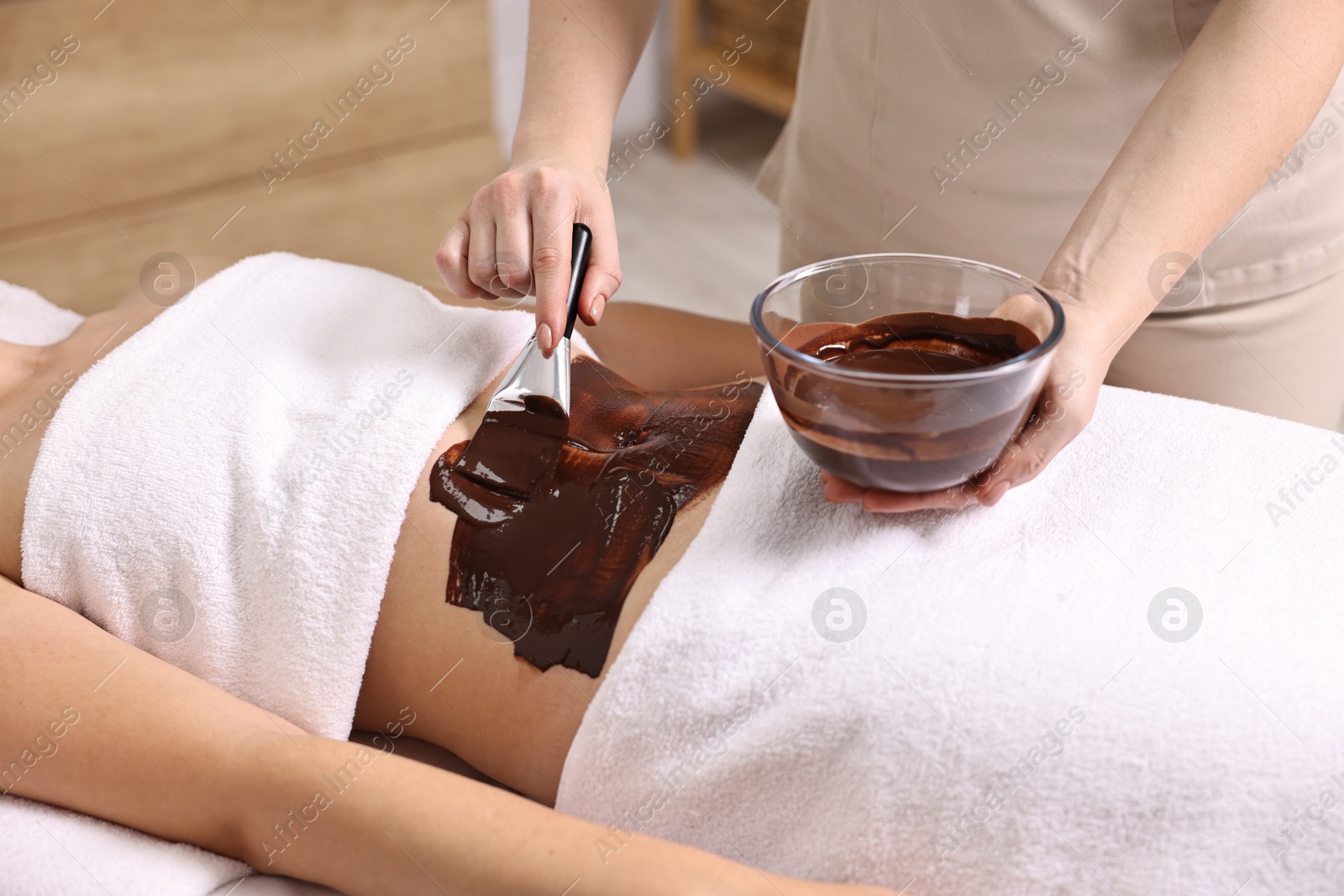 Photo of Chocolate body wrap. Spa worker applying mask onto woman's belly in salon, closeup