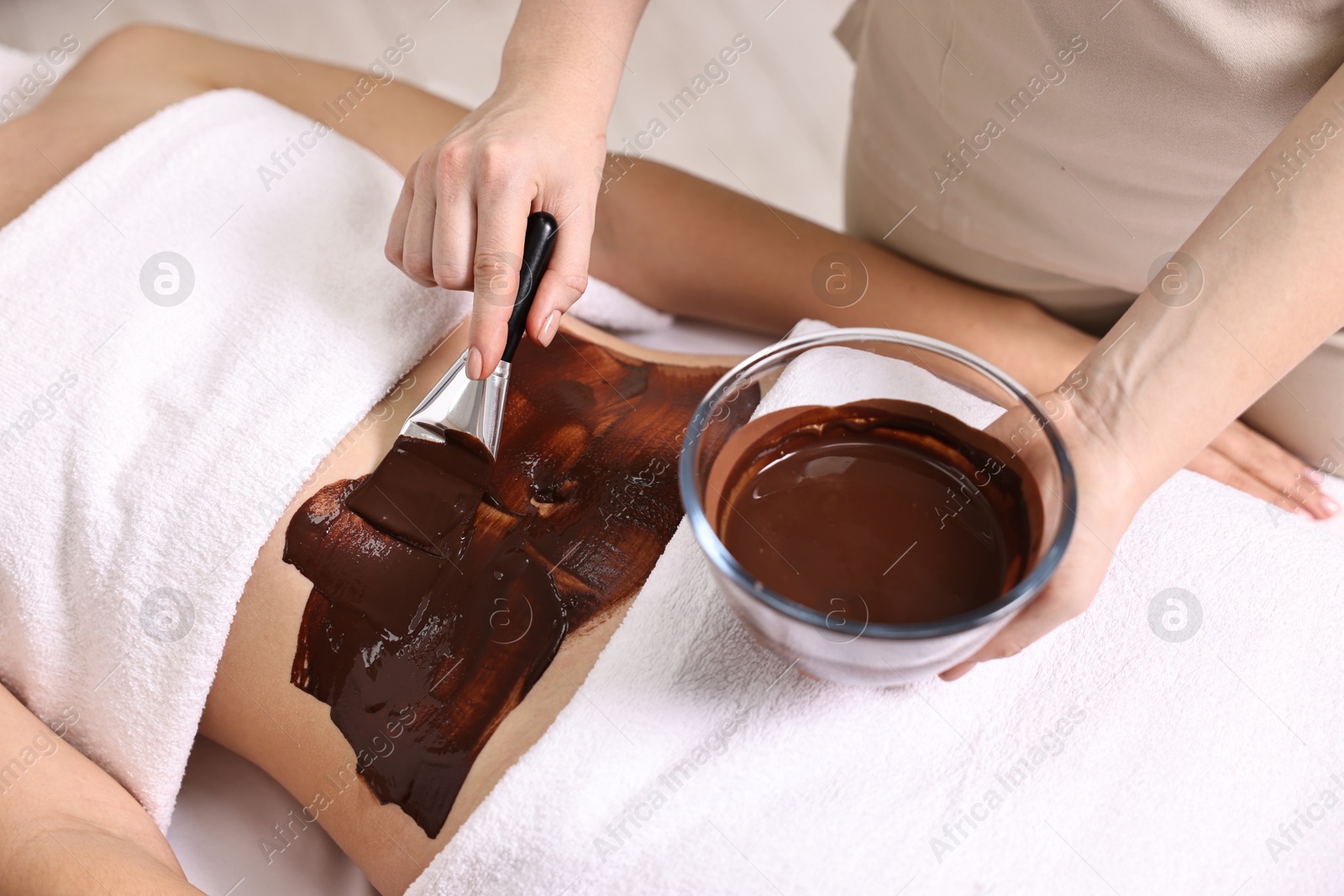 Photo of Chocolate body wrap. Spa worker applying mask onto woman's belly in salon, closeup