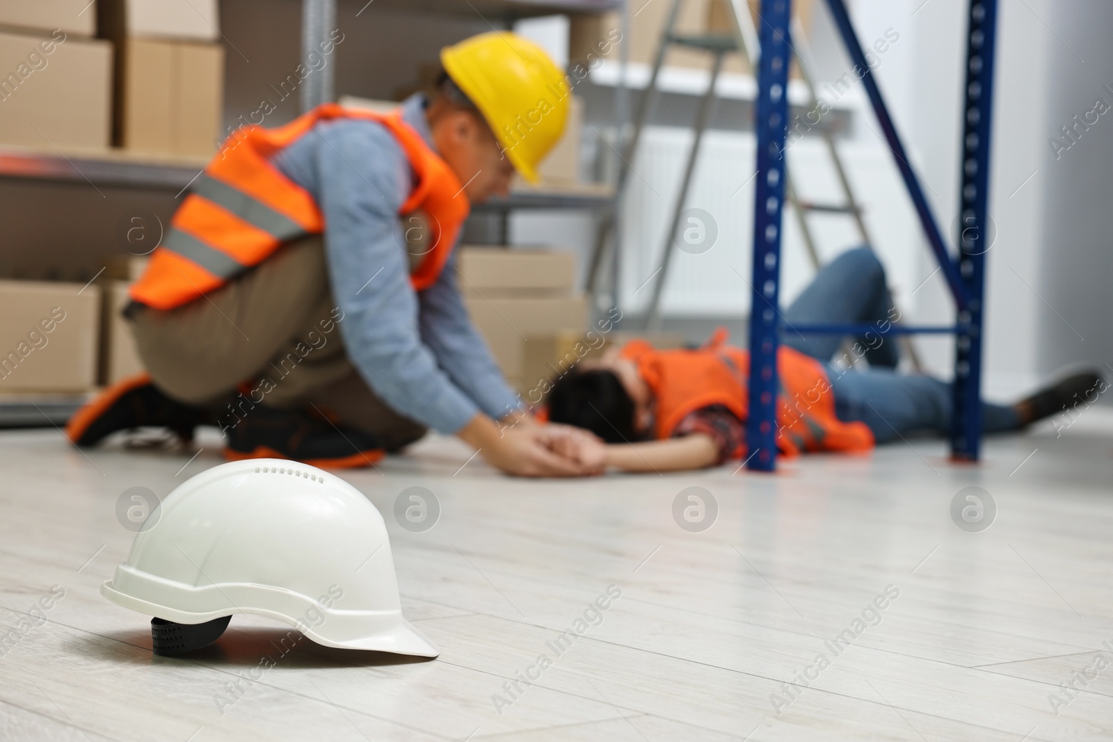 Photo of First aid. Hardhat on floor and man helping his unconscious colleague in warehouse, selective focus