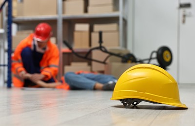 Photo of First aid. Hardhat on floor and man helping his unconscious colleague in warehouse, selective focus