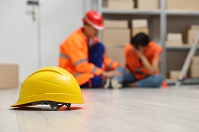 Photo of First aid. Hardhat on floor and man helping his injured colleague in warehouse, selective focus