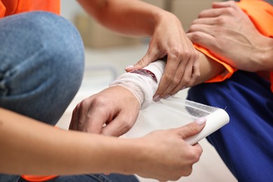 Photo of First aid. Woman putting bandage on her colleague's injured wrist after work accident indoors, closeup