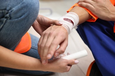 Photo of First aid. Woman putting bandage on her colleague's injured wrist after work accident indoors, closeup