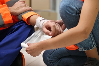 Photo of First aid. Woman putting bandage on her colleague's injured wrist after work accident indoors, closeup