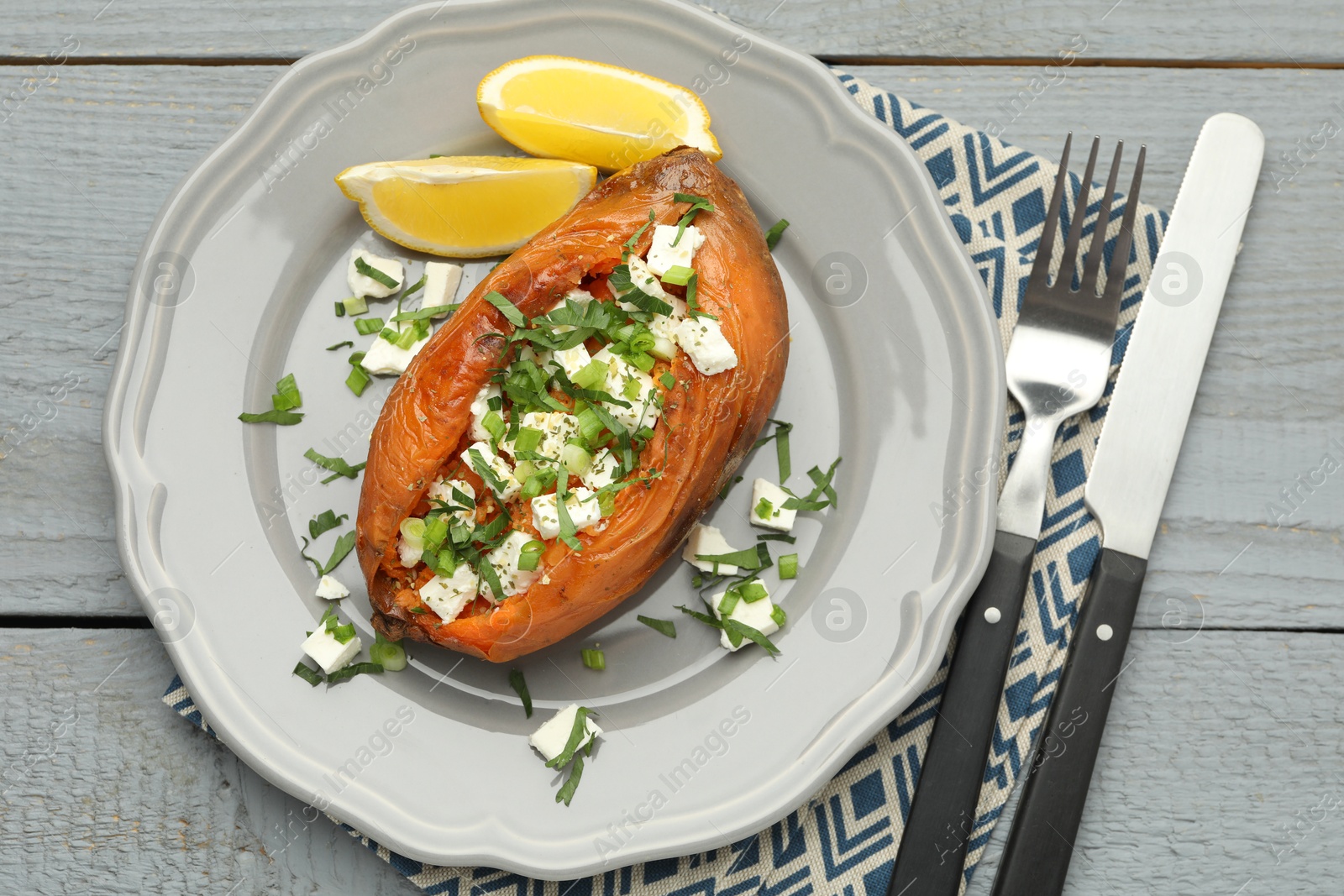 Photo of Tasty cooked sweet potato with feta cheese, lemon, green onion and cutlery on grey wooden table, top view