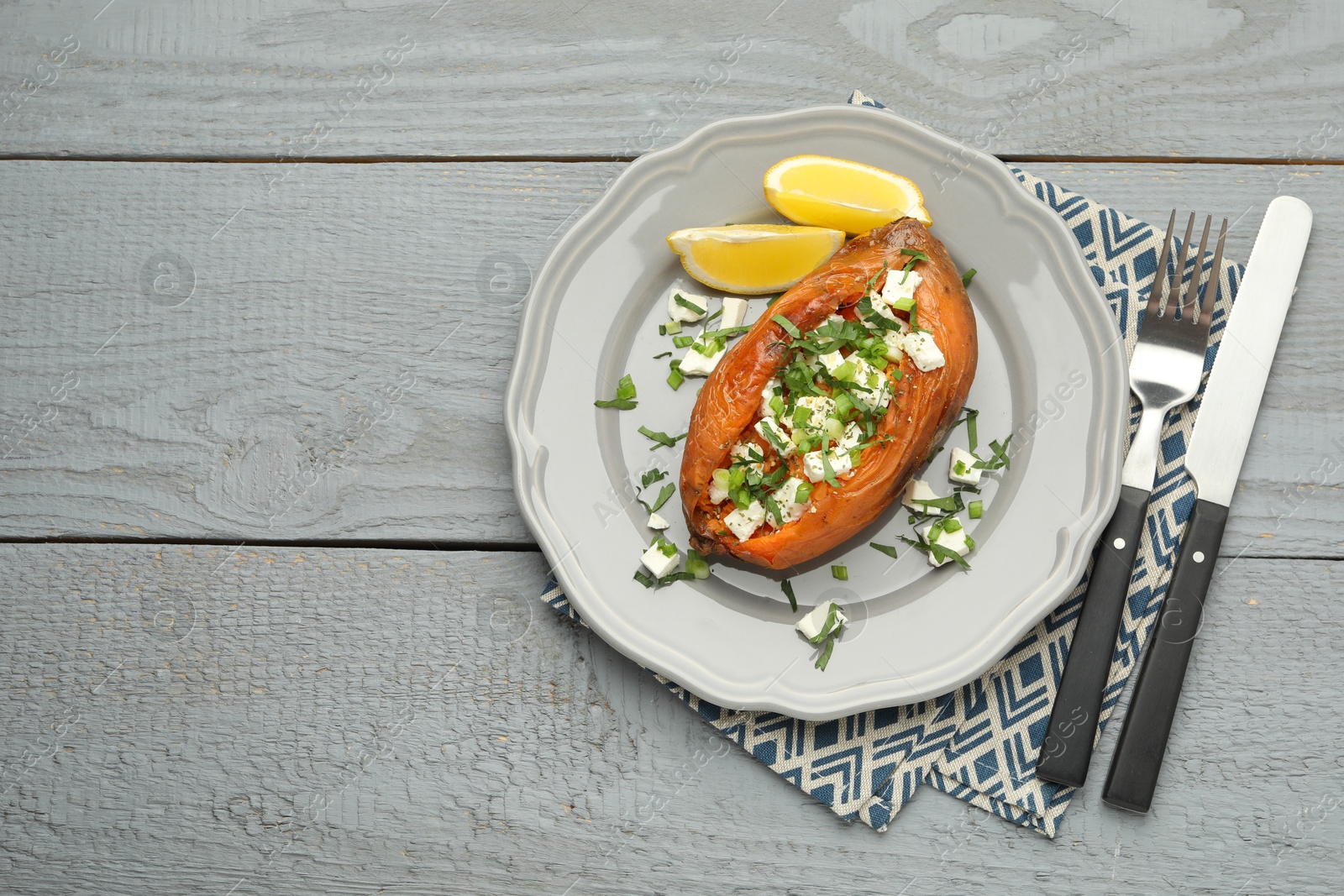 Photo of Tasty cooked sweet potato with feta cheese, lemon, green onion and cutlery on grey wooden table, top view. Space for text