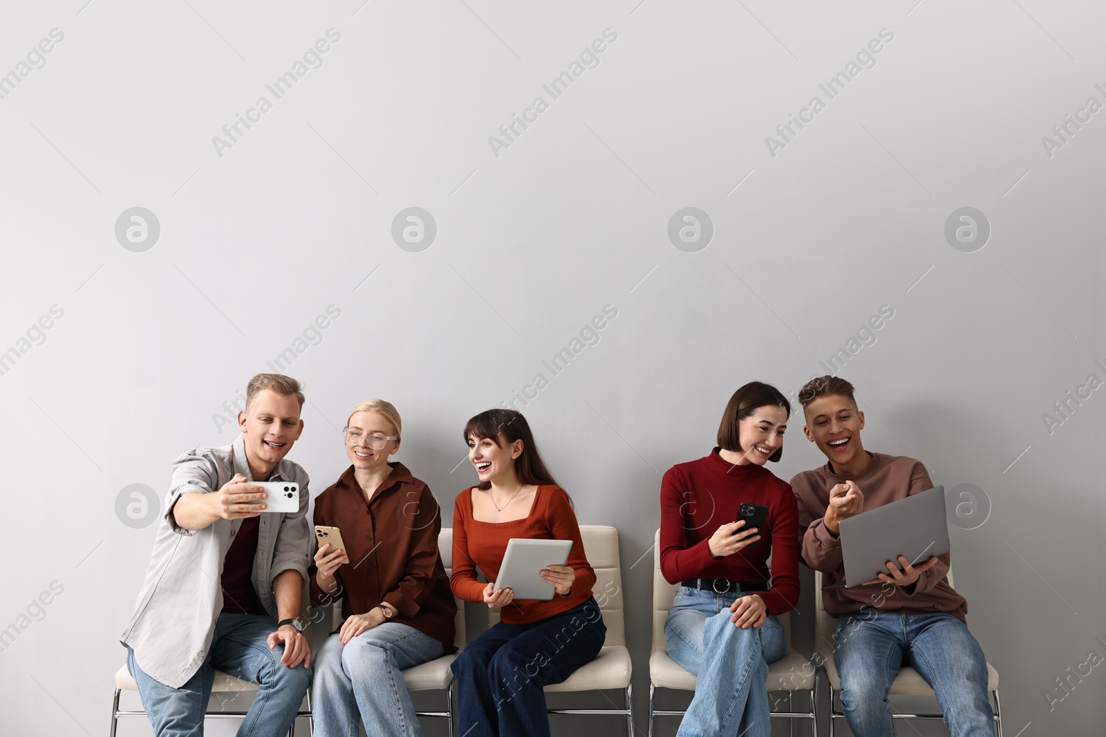 Photo of Group of people using different gadgets on chairs near light grey wall indoors. Modern technology