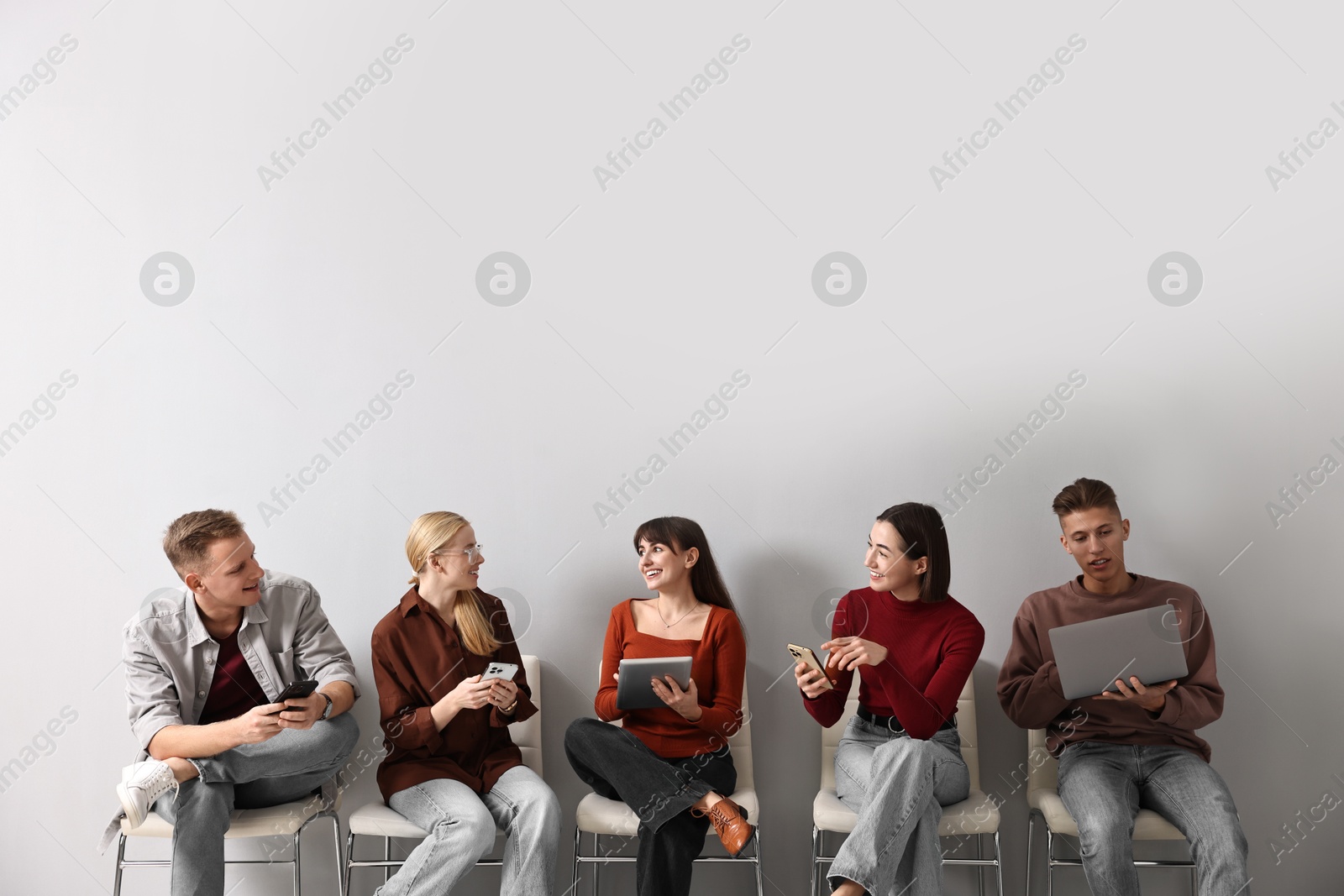 Photo of Group of people using different gadgets on chairs near light grey wall indoors. Modern technology