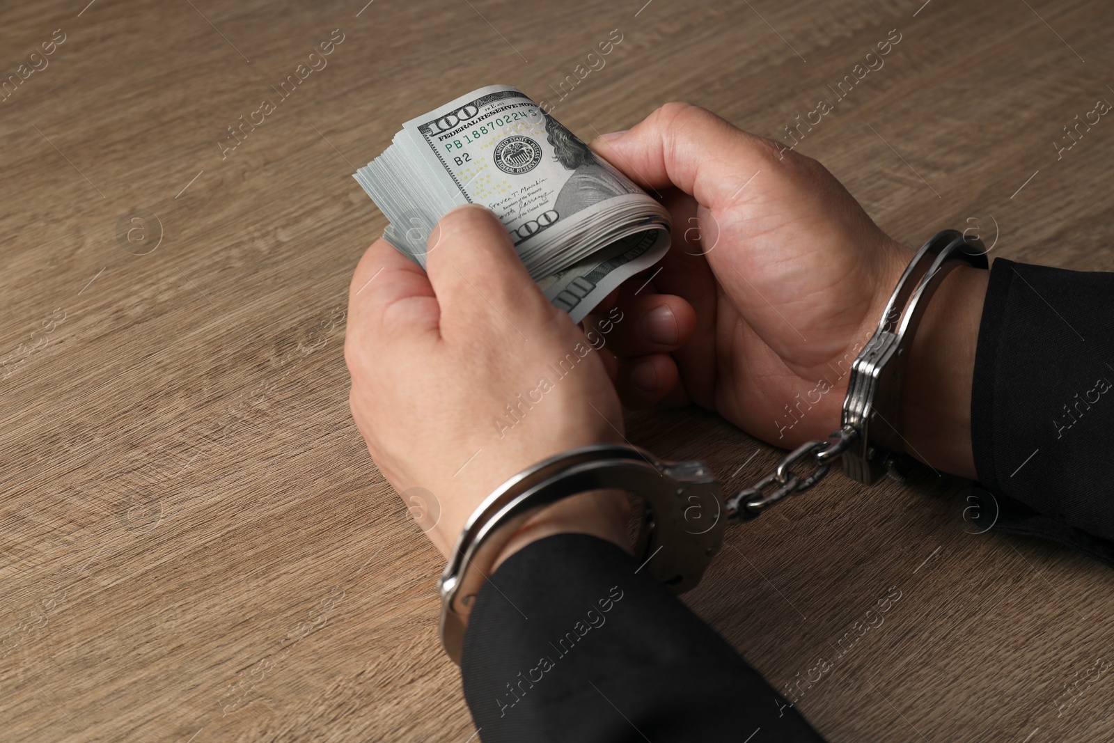 Photo of Corruption. Man in handcuffs holding dollar banknotes at wooden table, closeup