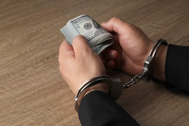 Photo of Corruption. Man in handcuffs holding dollar banknotes at wooden table, closeup