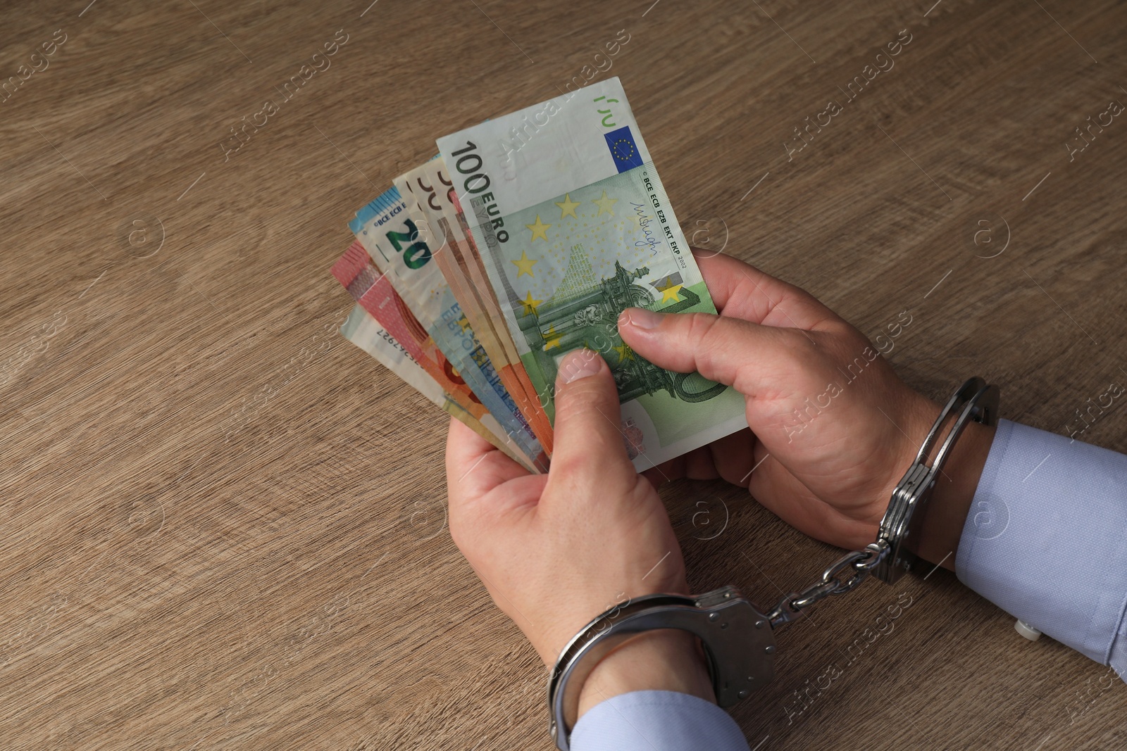 Photo of Corruption. Man in handcuffs holding euro banknotes at wooden table, closeup