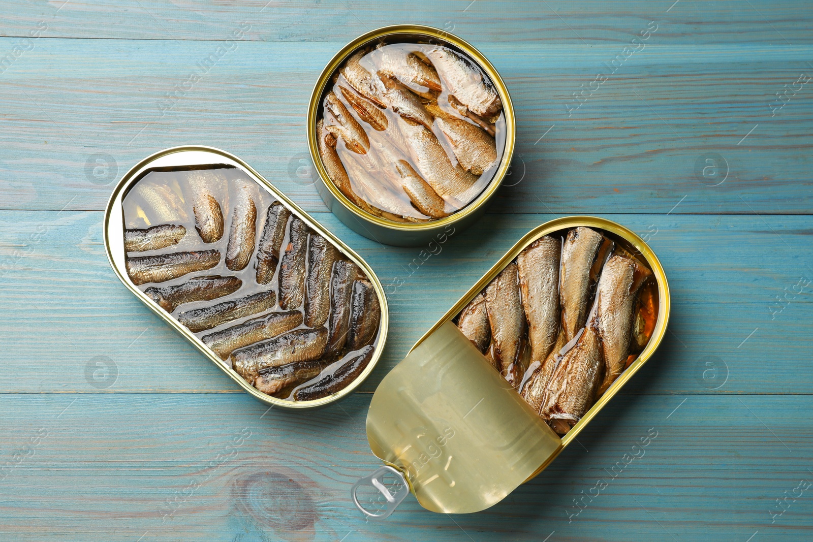 Photo of Tasty sprats in tin cans on light blue wooden table, flat lay