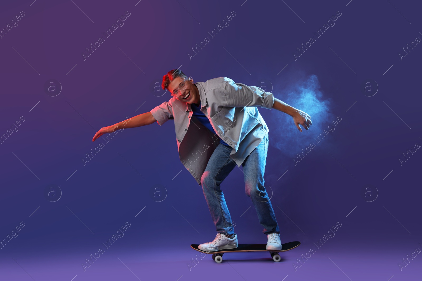 Photo of Happy man riding skateboard in smoke and color lights against blue background