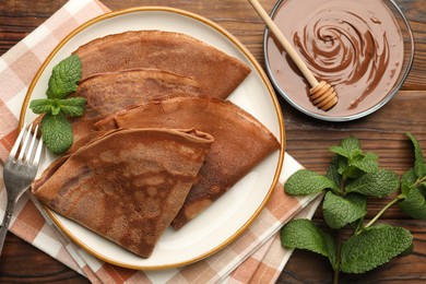 Photo of Delicious chocolate crepes, sweet sauce and mint on wooden table, flat lay