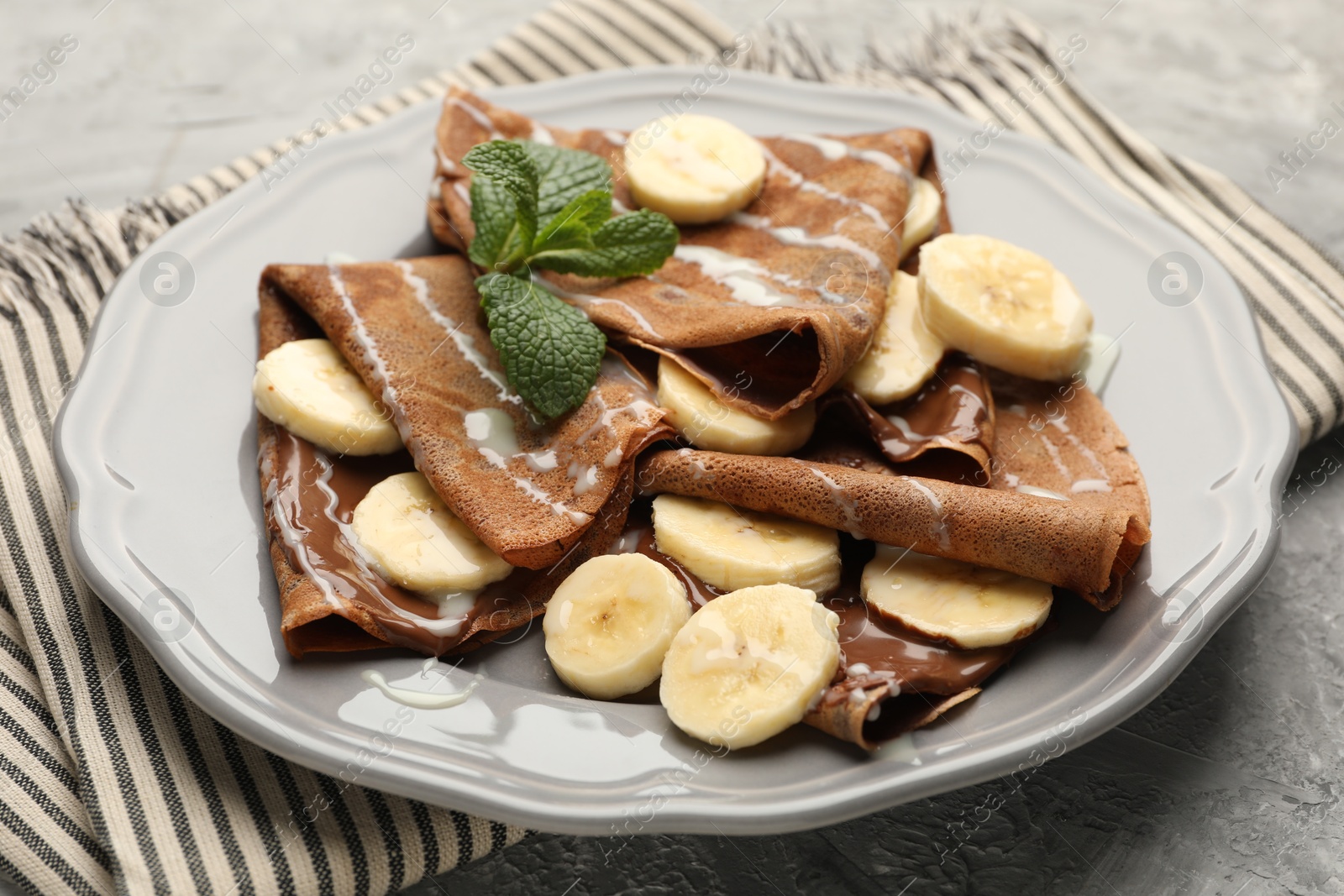 Photo of Delicious chocolate crepes with banana, condensed milk and mint served on grey table, closeup