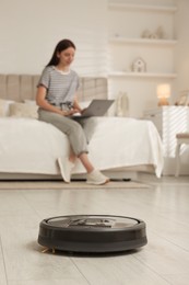 Photo of Young woman using laptop at home, focus on robotic vacuum cleaner