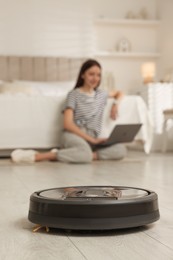 Photo of Young woman using laptop at home, focus on robotic vacuum cleaner