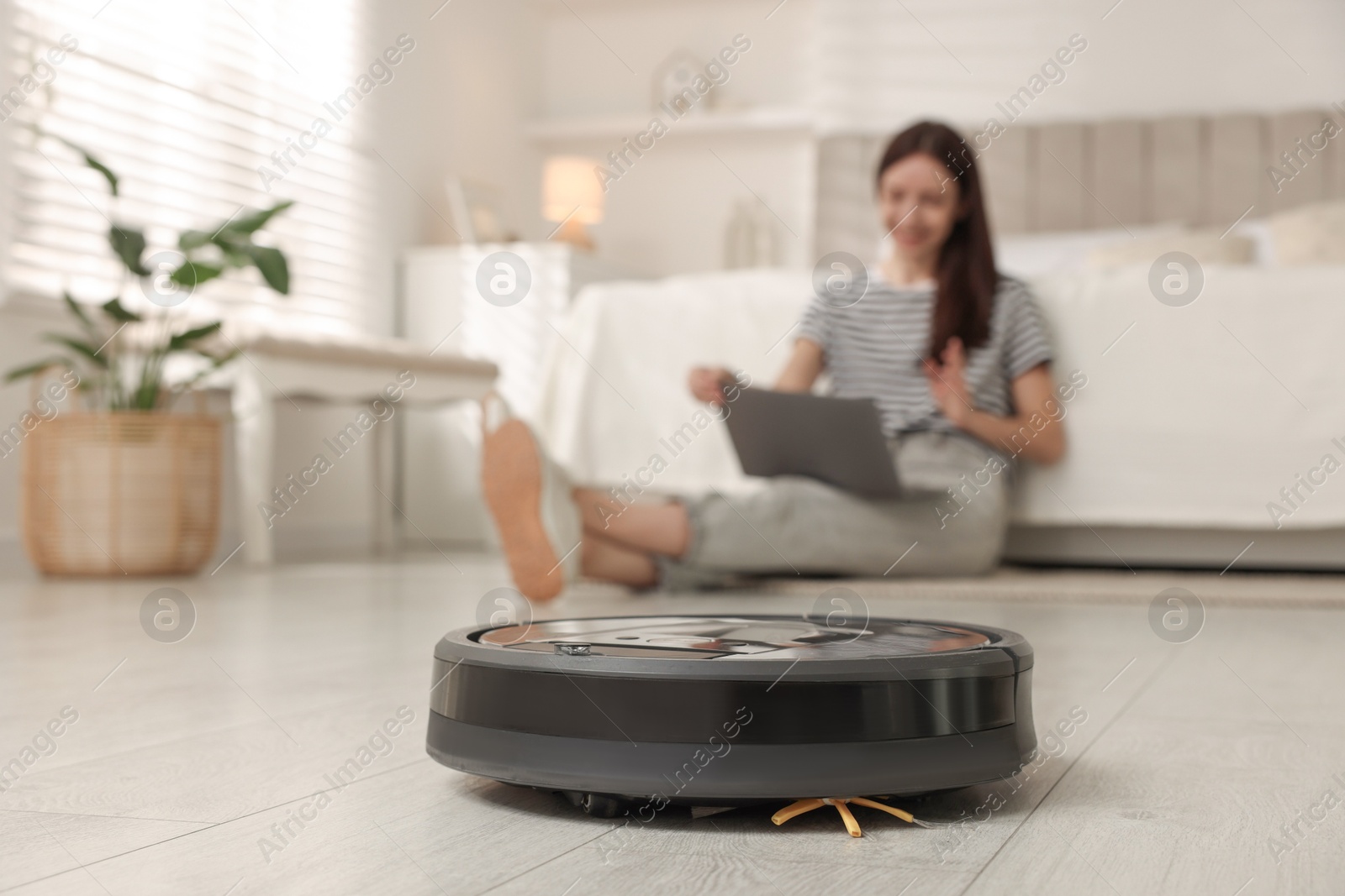 Photo of Young woman using laptop at home, focus on robotic vacuum cleaner