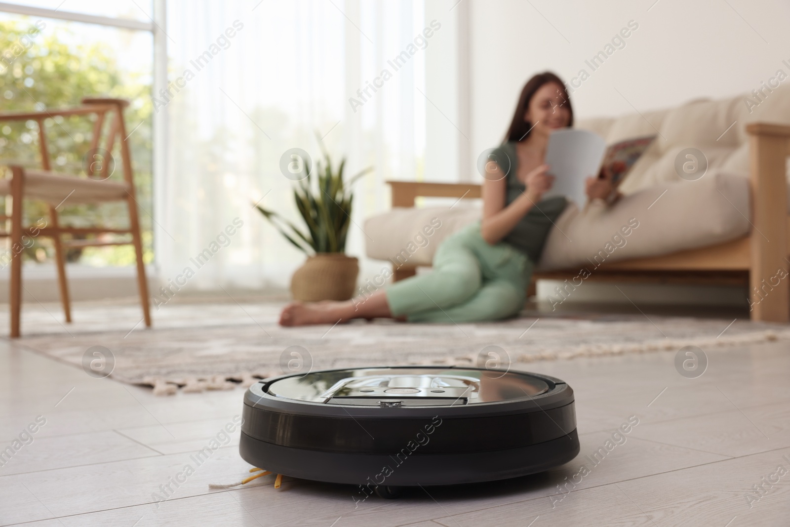 Photo of Young woman reading magazine at home, focus on robotic vacuum cleaner