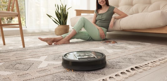 Photo of Young woman resting at home, focus on robotic vacuum cleaner