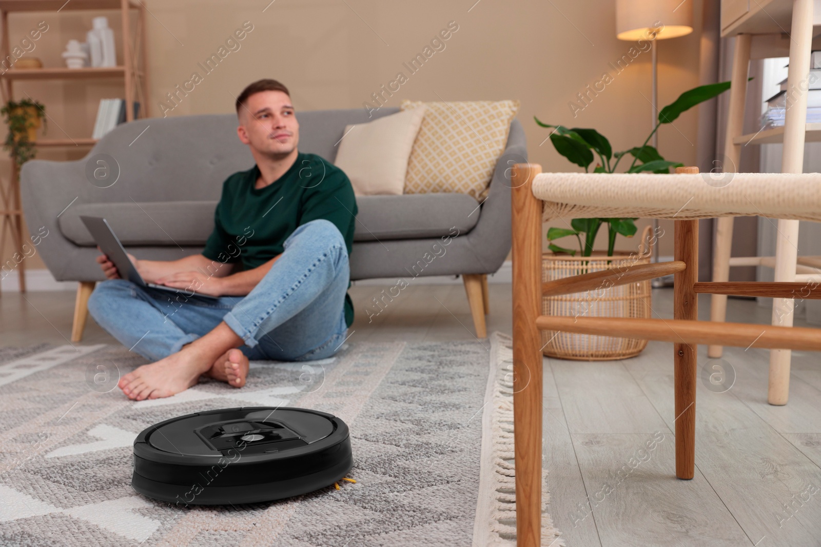 Photo of Young man using laptop at home, focus on robotic vacuum cleaner