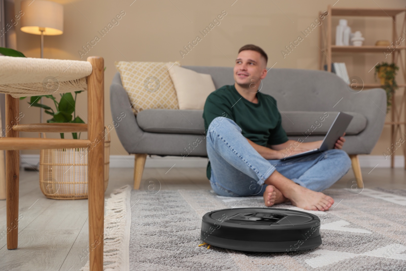 Photo of Young man using laptop at home, focus on robotic vacuum cleaner