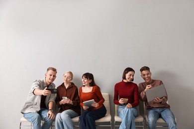 Group of people using different gadgets on chairs near light grey wall indoors. Modern technology