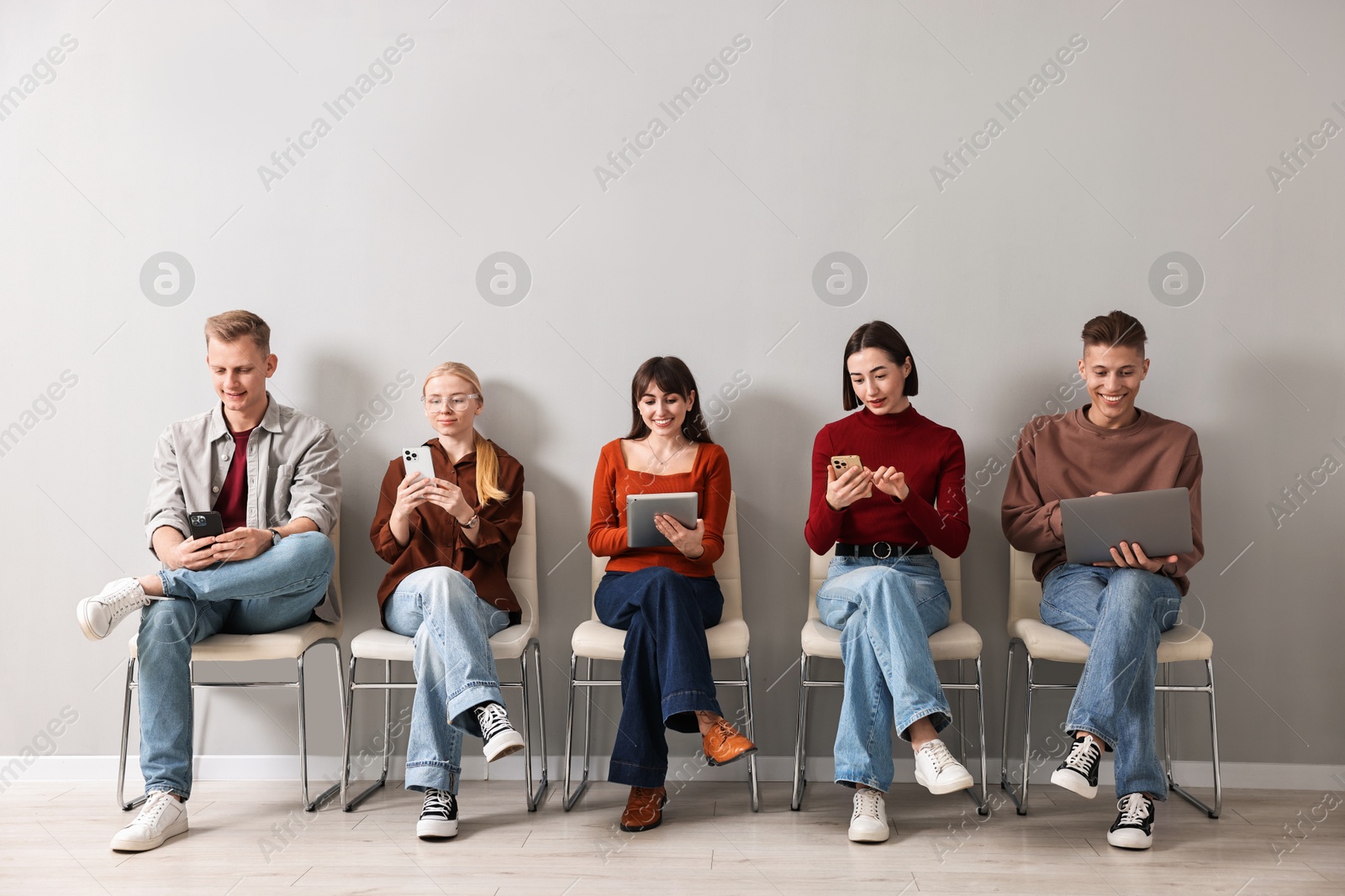 Photo of Group of people using different gadgets on chairs near light grey wall indoors. Modern technology