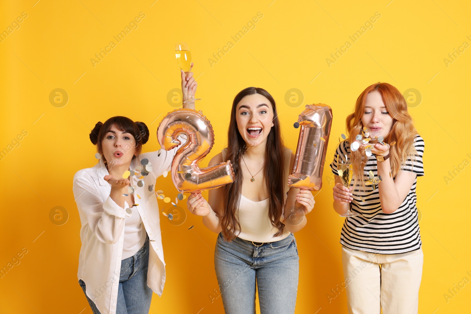 Photo of Coming of age party - 21st birthday. Group of young women celebrating with number shaped balloons and blowing confetti on yellow background