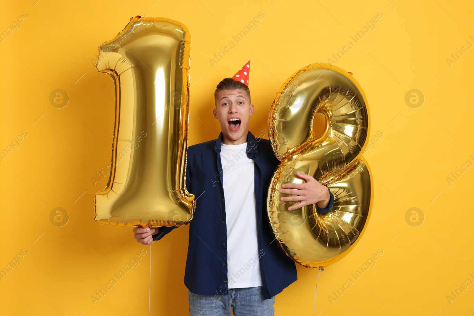 Photo of Coming of age party - 18th birthday. Emotional young man with number shaped balloons and hat on yellow background