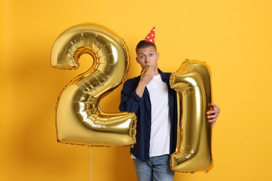 Photo of Coming of age party - 21st birthday. Happy young man with number shaped balloons, blower and hat on yellow background