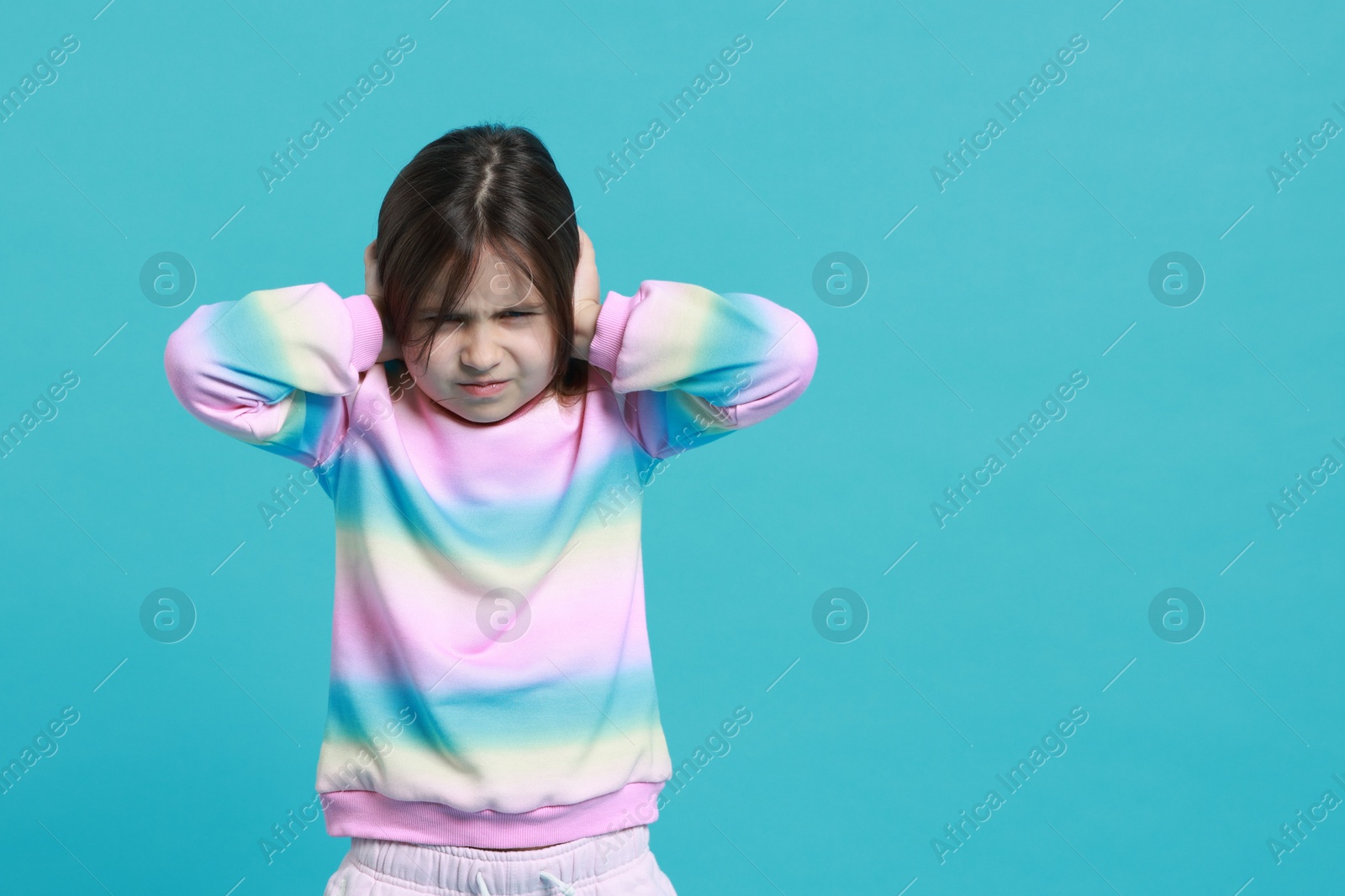 Photo of Annoyed little girl covering her ears from loud sound on light blue background. Space for text