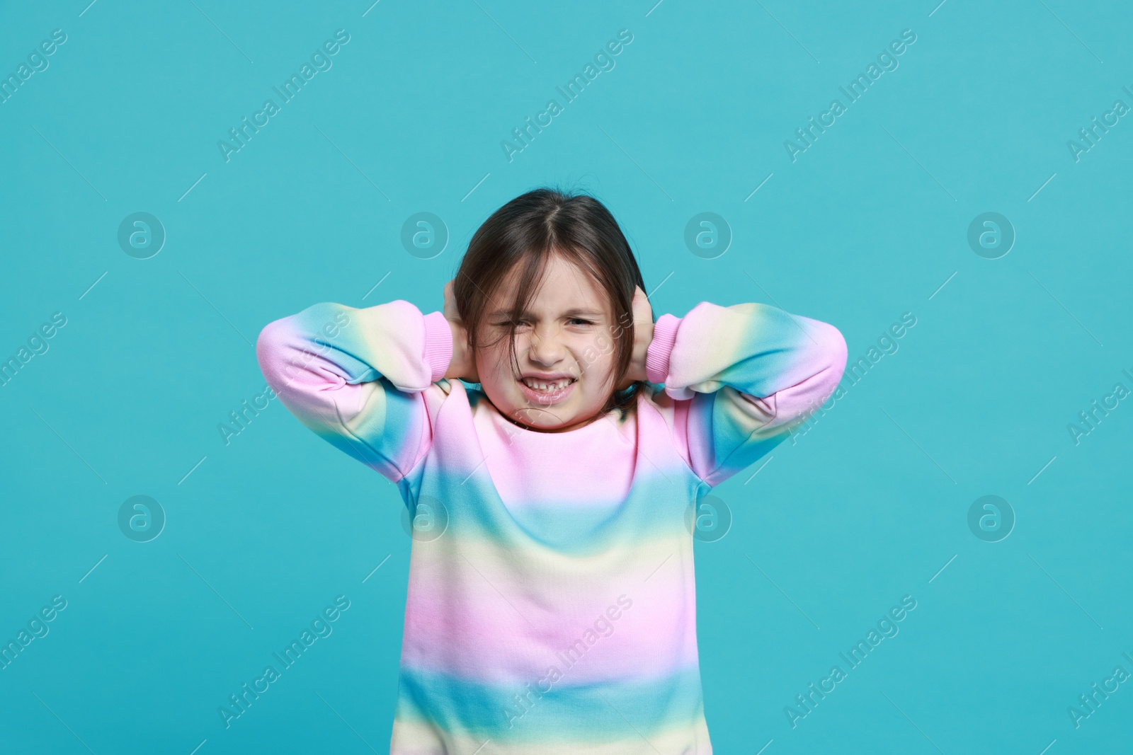 Photo of Annoyed little girl covering her ears from loud sound on light blue background