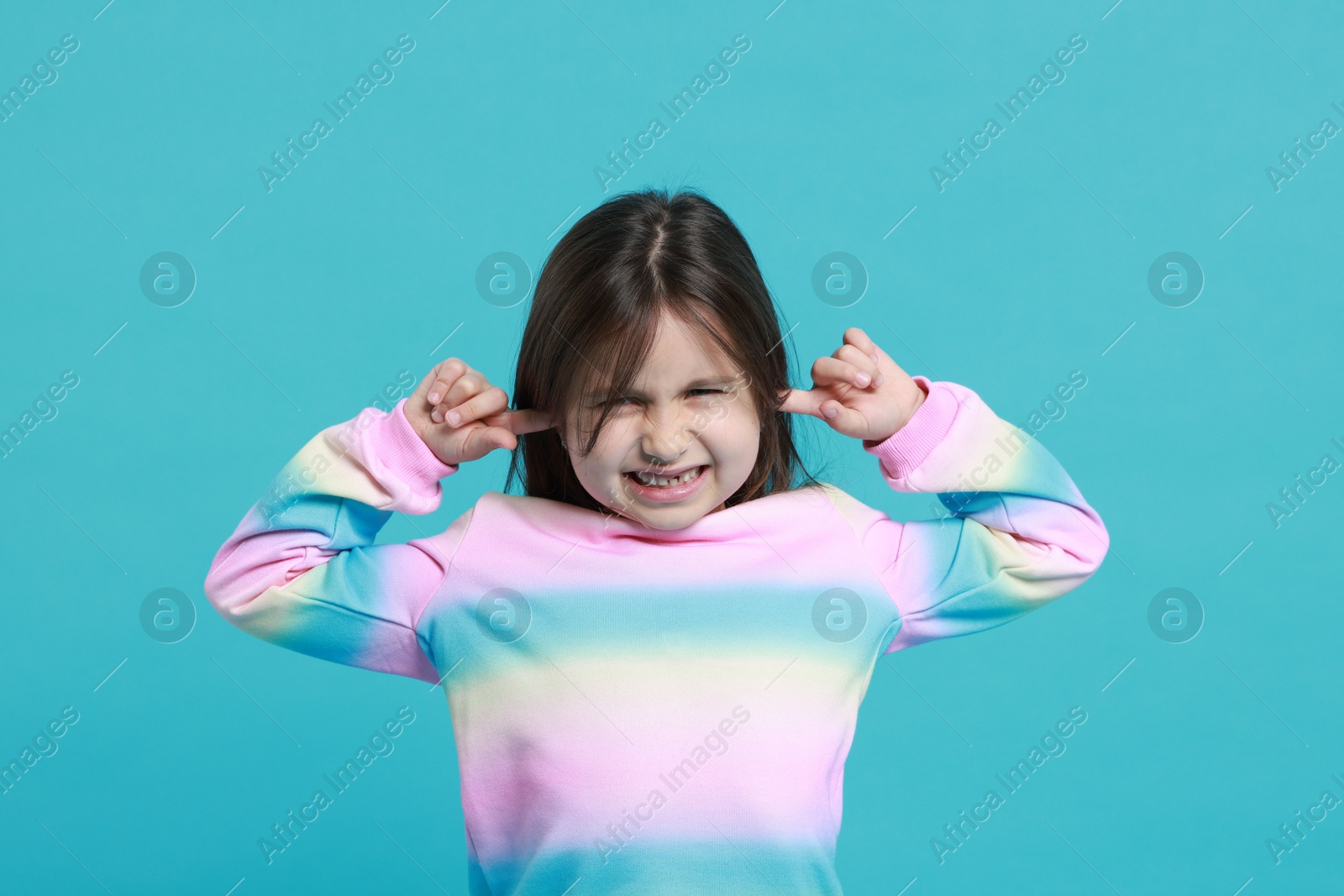 Photo of Annoyed little girl covering her ears from loud sound on light blue background