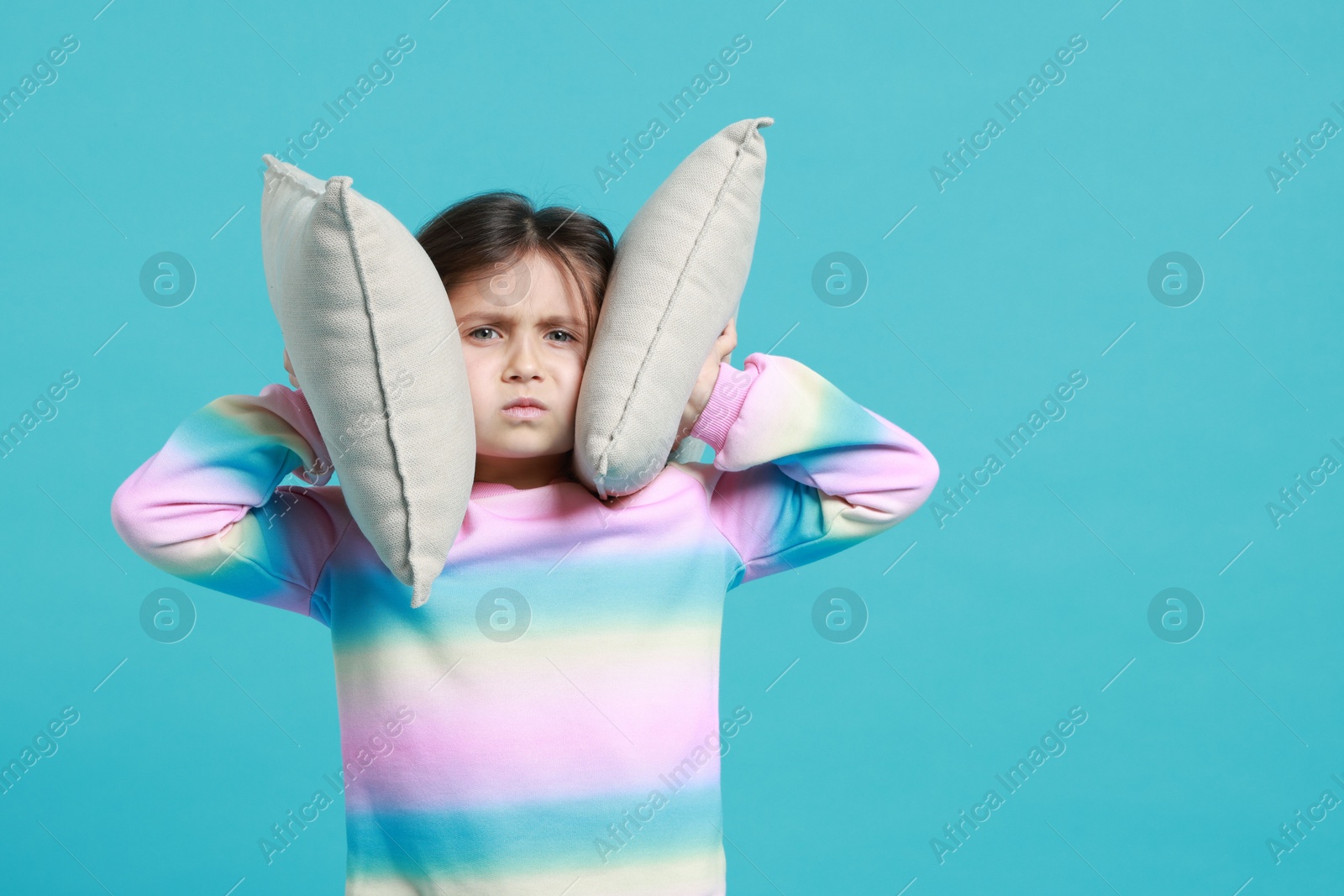 Photo of Annoyed little girl covering her ears with pillows from loud sound on light blue background. Space for text