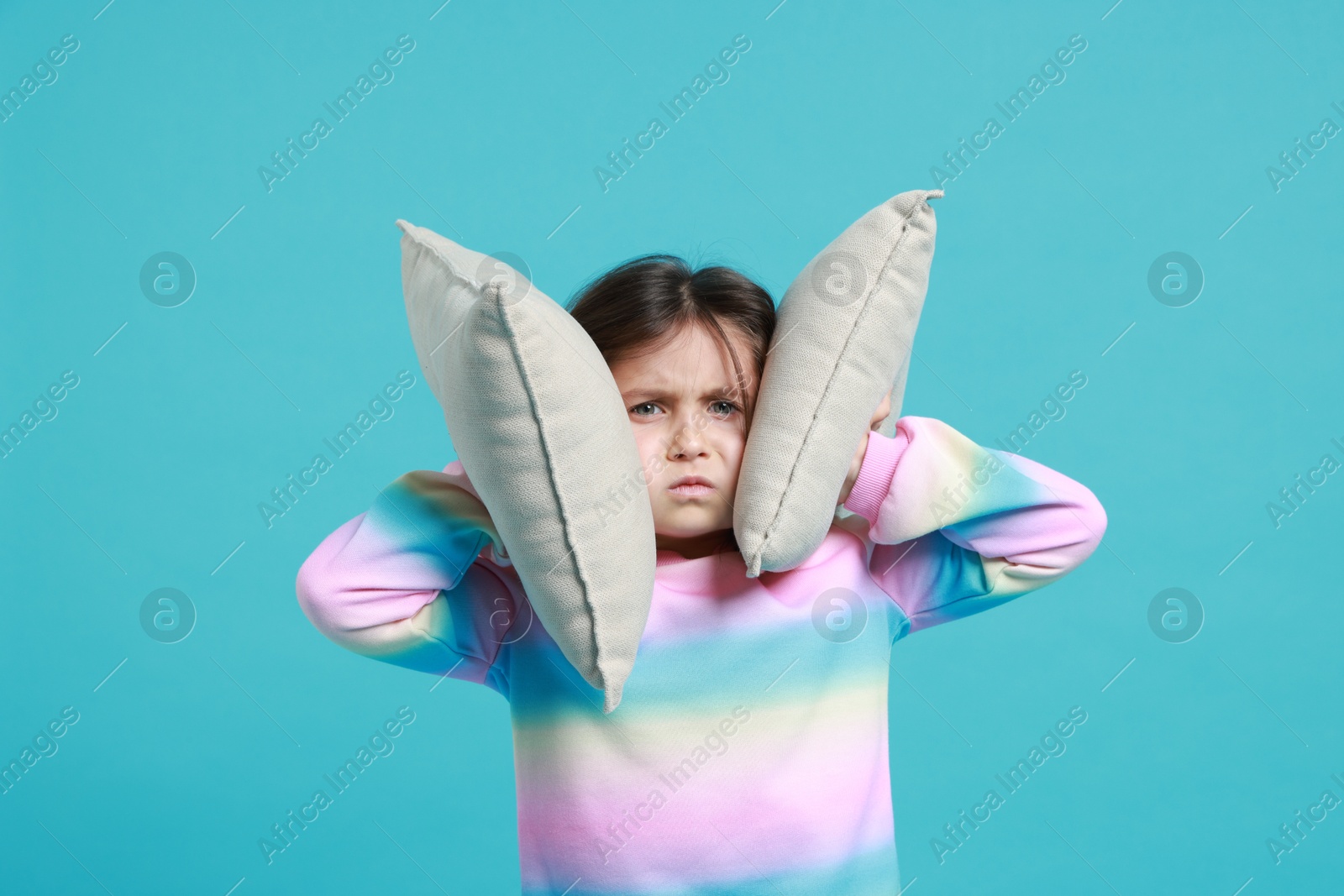 Photo of Annoyed little girl covering her ears with pillows from loud sound on light blue background