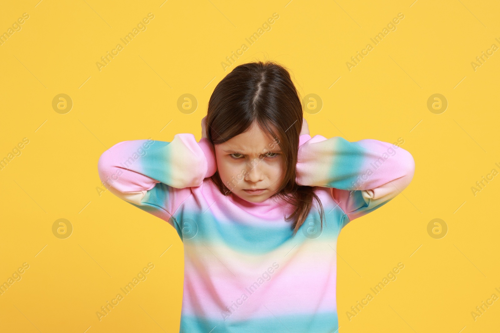 Photo of Annoyed little girl covering her ears from loud sound on yellow background