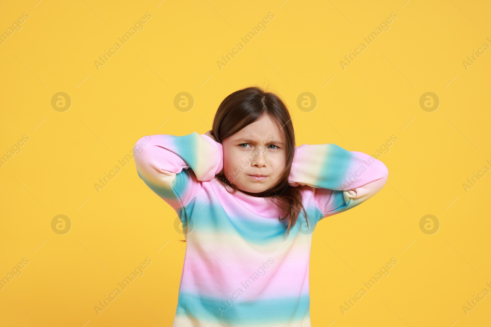 Photo of Annoyed little girl covering her ears from loud sound on yellow background