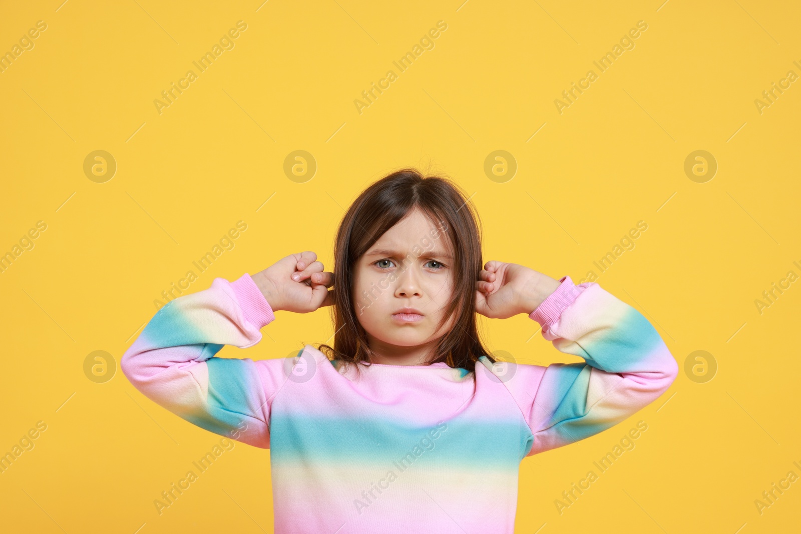 Photo of Annoyed little girl covering her ears from loud sound on yellow background