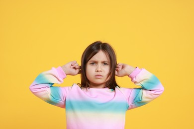 Photo of Annoyed little girl covering her ears from loud sound on yellow background