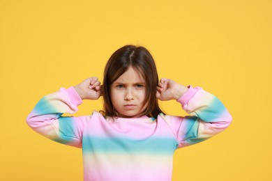 Photo of Annoyed little girl covering her ears from loud sound on yellow background