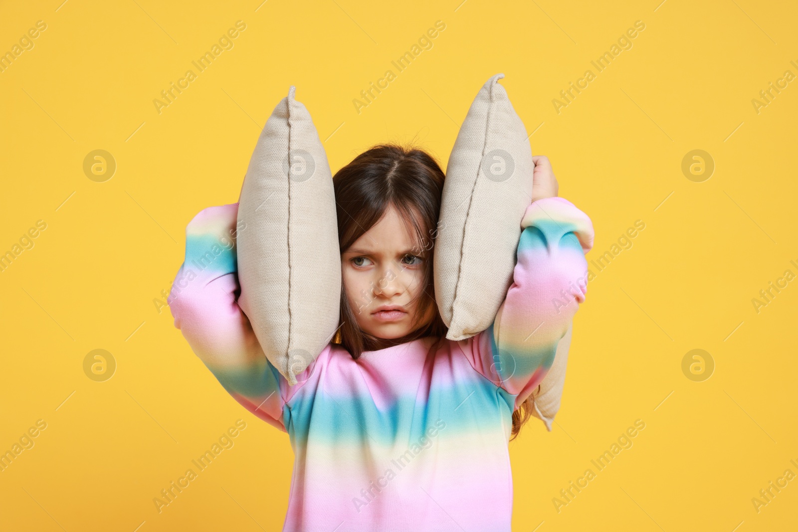 Photo of Annoyed little girl covering her ears with pillows from loud sound on yellow background