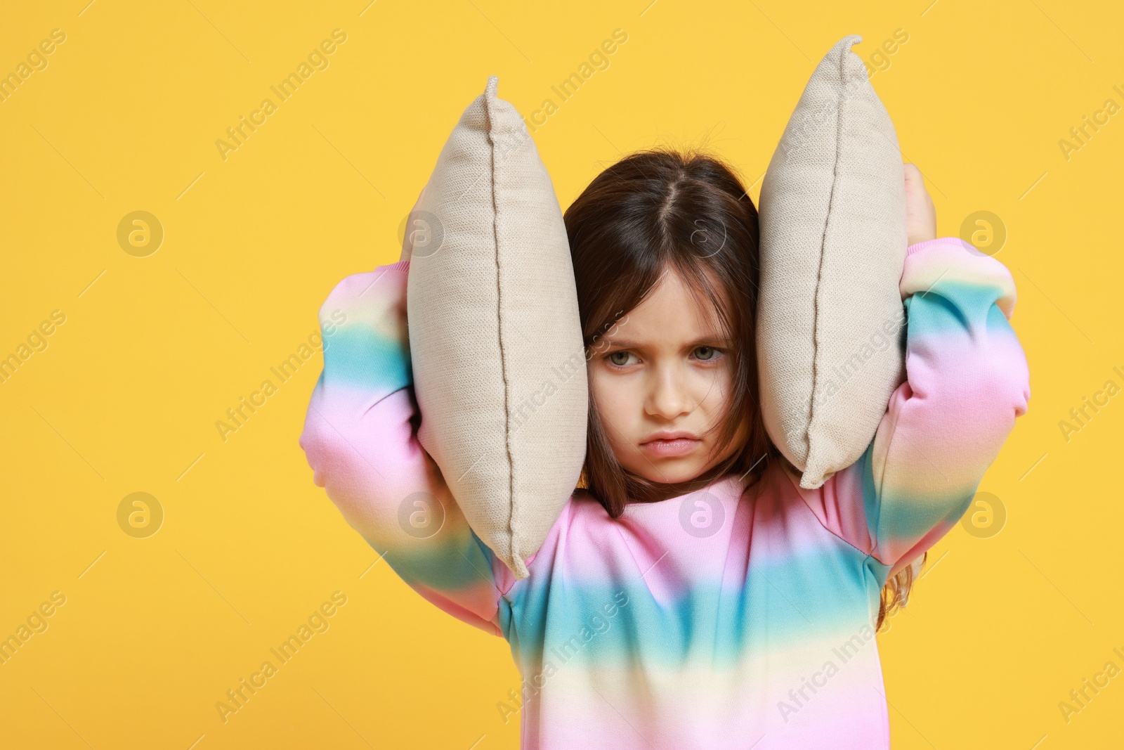 Photo of Exhausted little girl covering her ears with pillows from loud sound on yellow background. Space for text