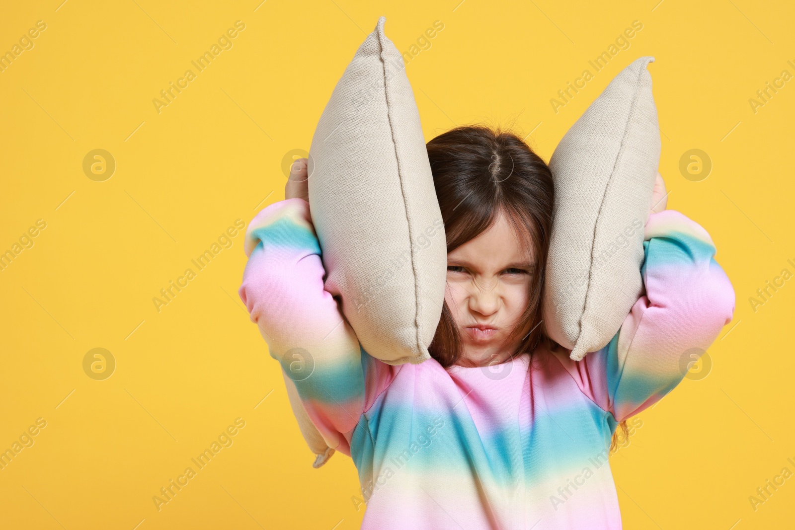 Photo of Annoyed little girl covering her ears with pillows from loud sound on yellow background. Space for text