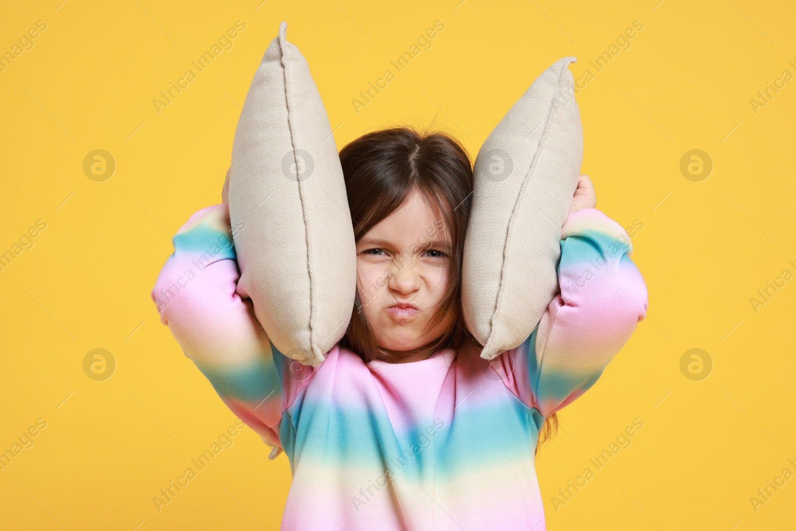 Photo of Annoyed little girl covering her ears with pillows from loud sound on yellow background