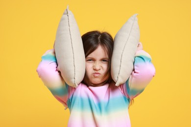 Photo of Annoyed little girl covering her ears with pillows from loud sound on yellow background